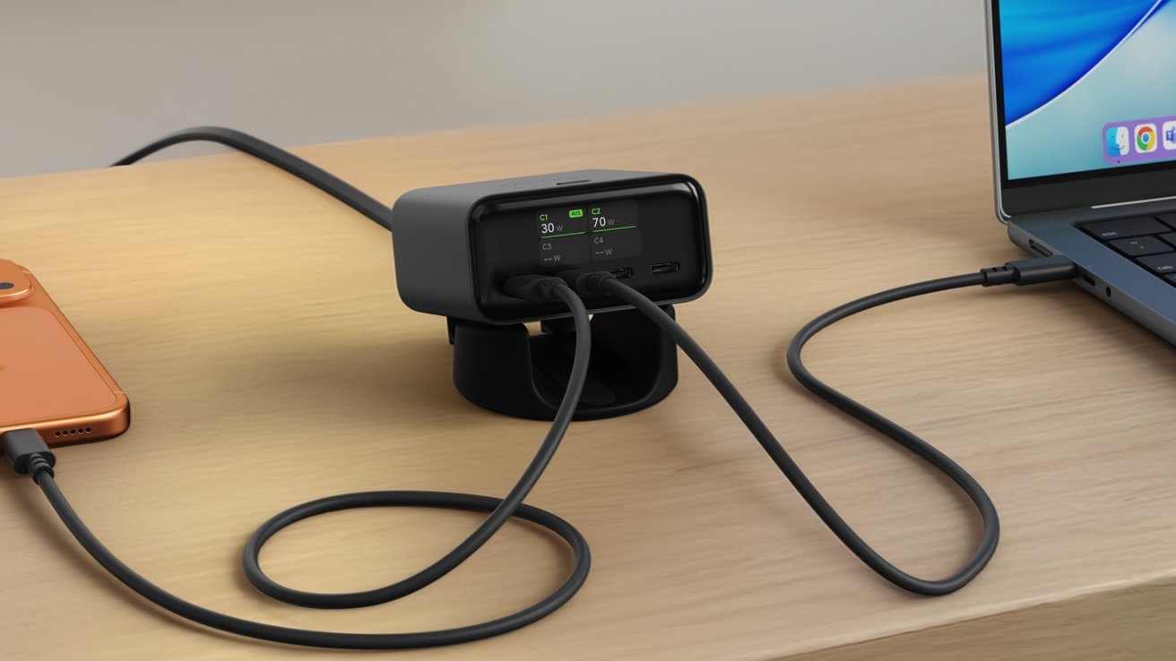 Compact black charging hub on a wooden desk, with cables connected to an orange smartphone and a laptop, showing digital wattage readings on a small front display