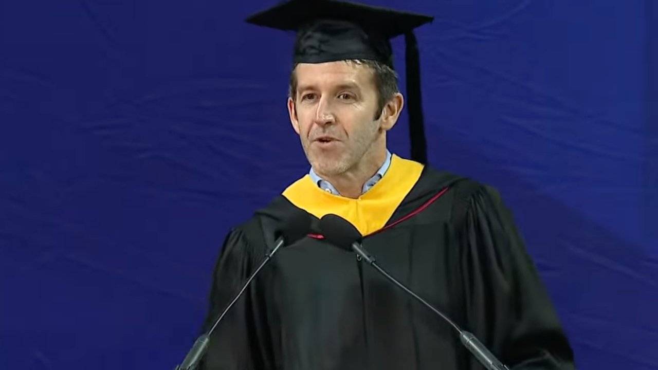 Man in black graduation cap and gown with yellow hood speaks at podium with two microphones against a solid blue backdrop