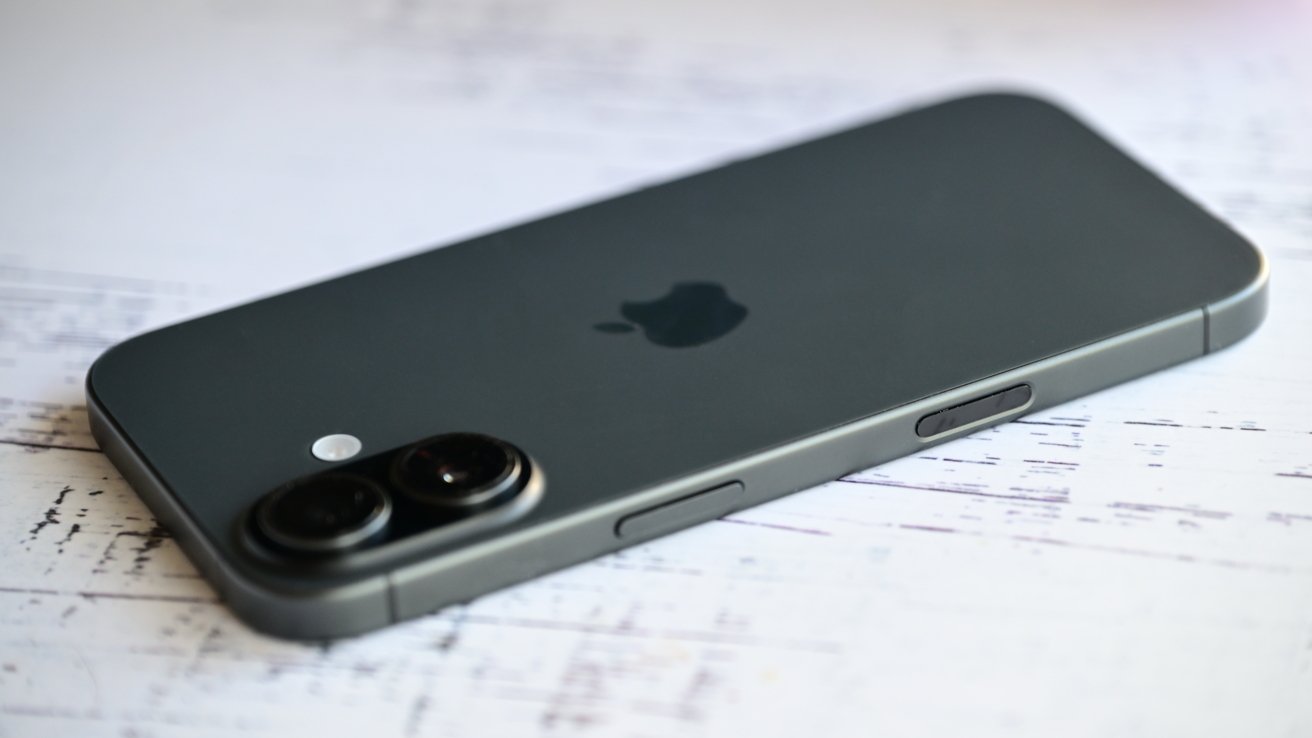 Dark gray iPhone lying face down on a light wooden surface, showing dual rear cameras, a small flash, side buttons, and centered Apple logo on the back