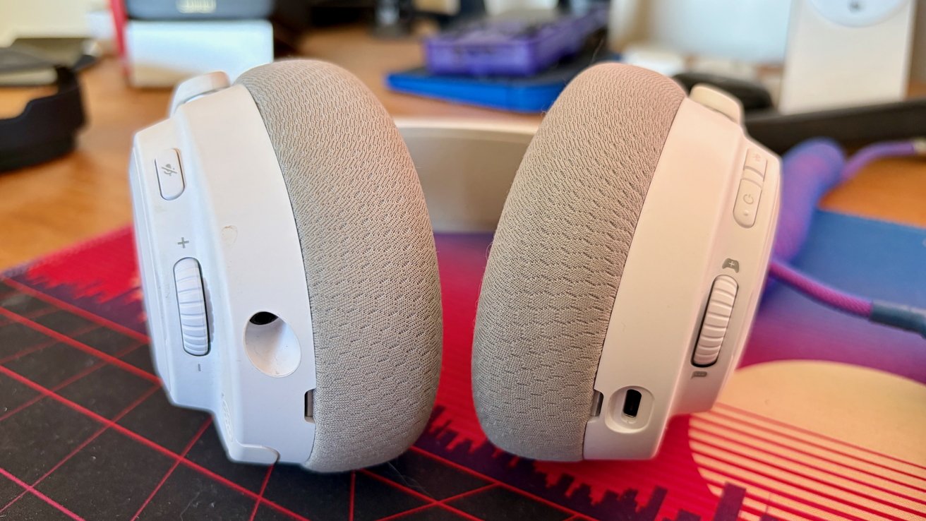 Close-up of white over-ear headphones with gray fabric ear cushions, showing volume rollers, buttons, and ports, resting on a colorful desk mat in a cluttered workspace background