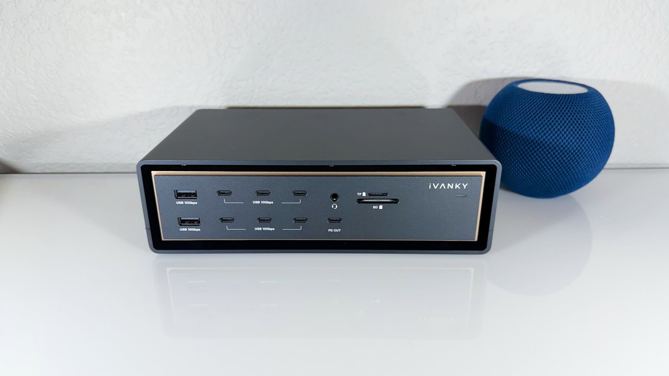 Black iVANKY docking station with multiple USB ports and SD card slots on a white desk, next to a small blue spherical smart speaker against a light textured wall