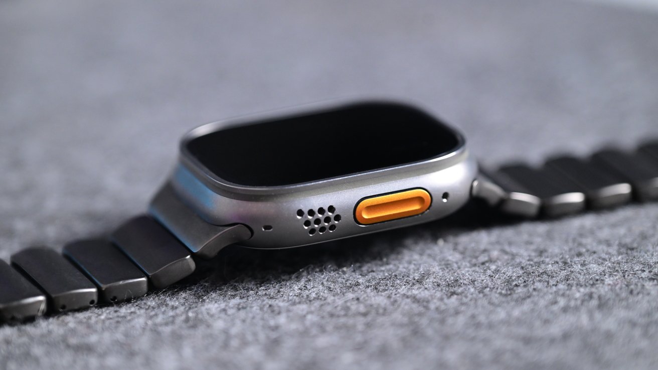Close-up of a modern smartwatch lying on its side, showing a silver case, bright orange side button, small speaker holes, and a dark metal link band on a soft gray surface