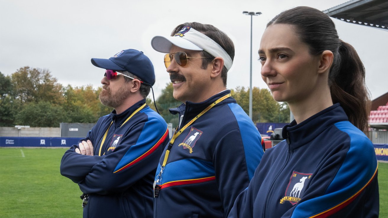 Three soccer coaches in matching navy tracksuits stand side by side on a field, watching intently, with stadium seating, green grass, and overcast sky in the background