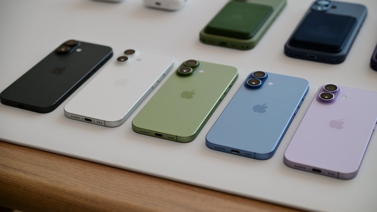 Row of colorful iPhones on a table, including black, white, green, blue, and purple models, all placed face down showing rear cameras and Apple logo