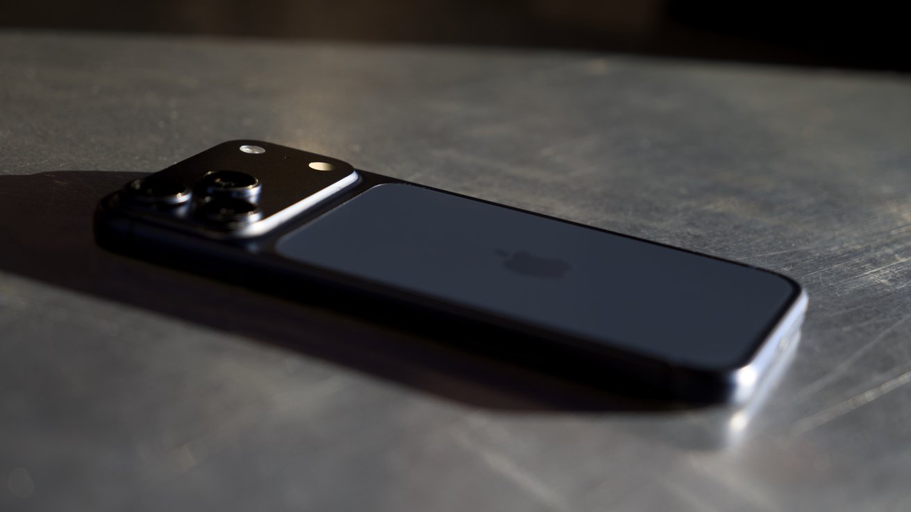 iPhone 17 Pro Max lying face down on a metallic surface, showing a raised triple camera bump and subtle reflections from low, directional lighting