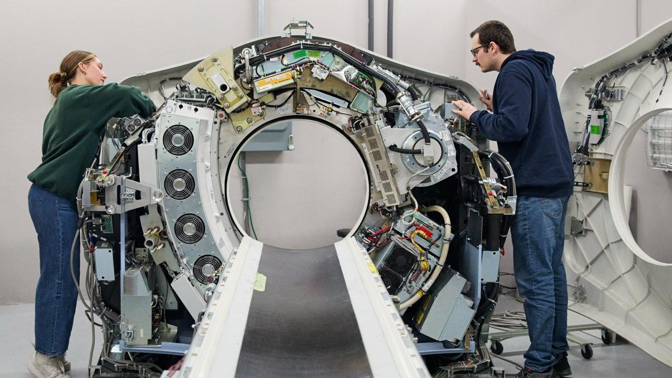 Two technicians stand on either side of an open CT scanner, working on exposed wires, fans, and electronic components around the circular opening and patient table.