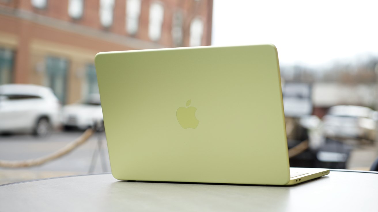 Light green Apple laptop open on an outdoor table, viewed from behind, with blurred cars and brick buildings in the background