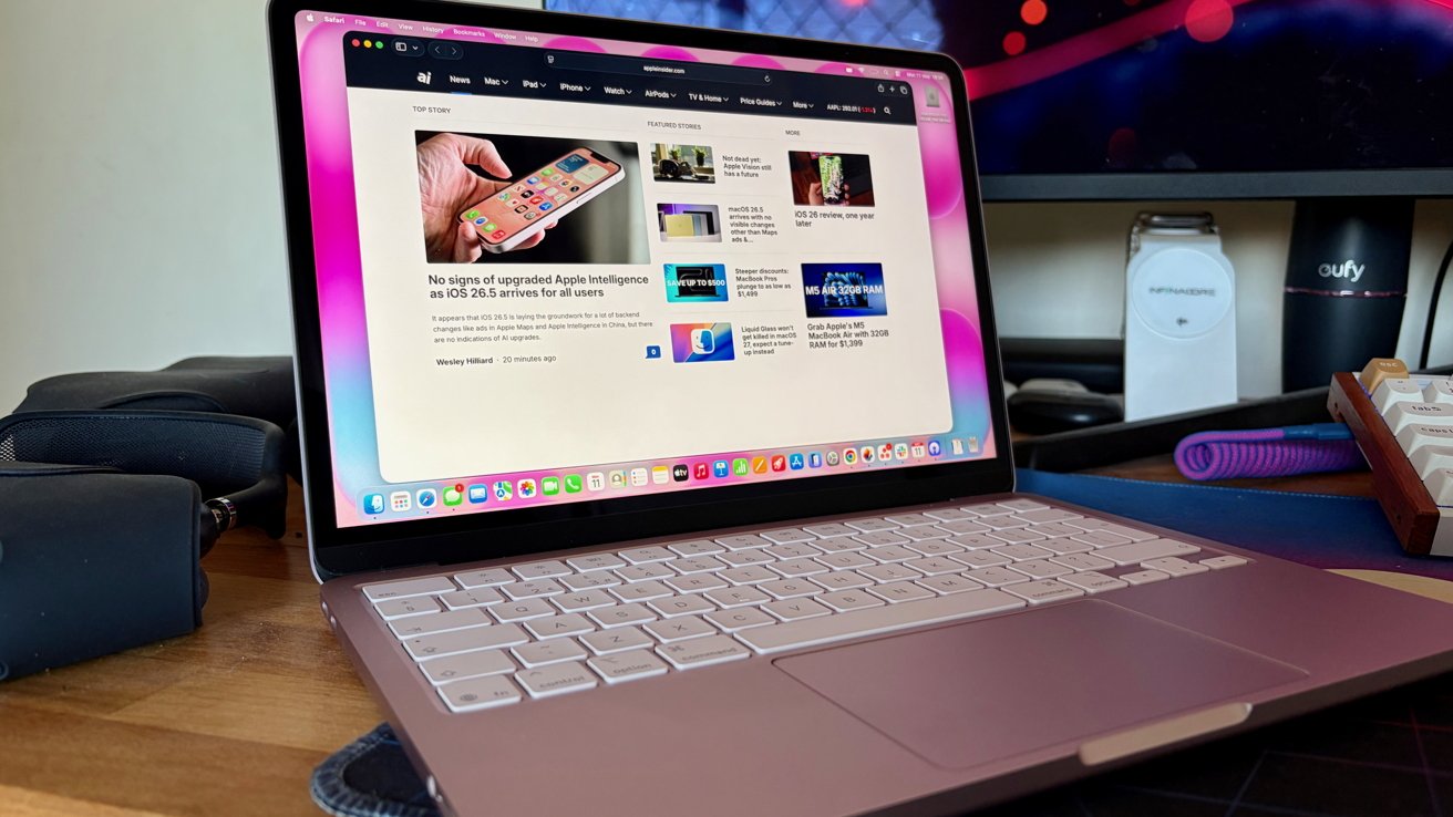Open pink MacBook on a wooden desk displaying a tech news website, with external monitor, keyboard, and various gadgets in the background of a tidy home office setup