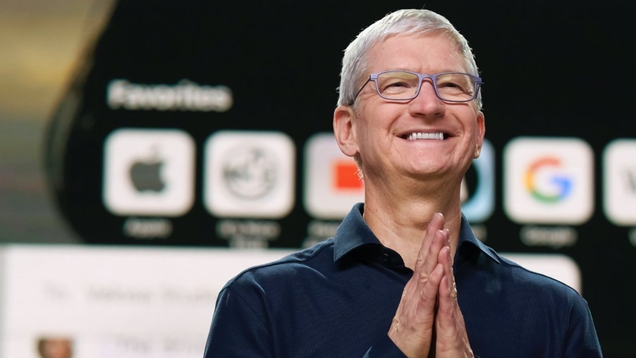 Smiling older man with short gray hair and glasses stands clasping his hands together, wearing a dark shirt, in front of a blurred screen showing large colorful app icons