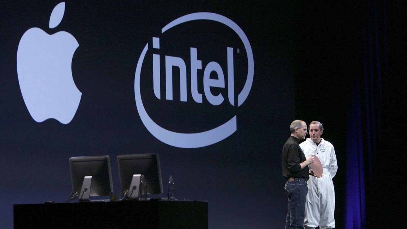 Two men on a dark stage beneath large Apple and Intel logos, talking beside two desktop computers; one man wears casual clothes, the other an Intel-branded white cleanroom suit holding a wafer