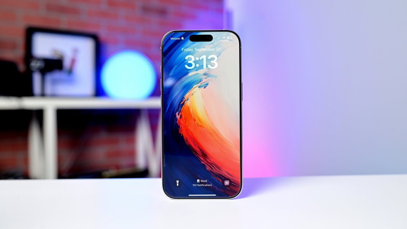 Modern smartphone standing upright on a white table, displaying 3:13 and colorful abstract wallpaper, with a blurred background of brick wall, shelves, and soft blue and pink lighting