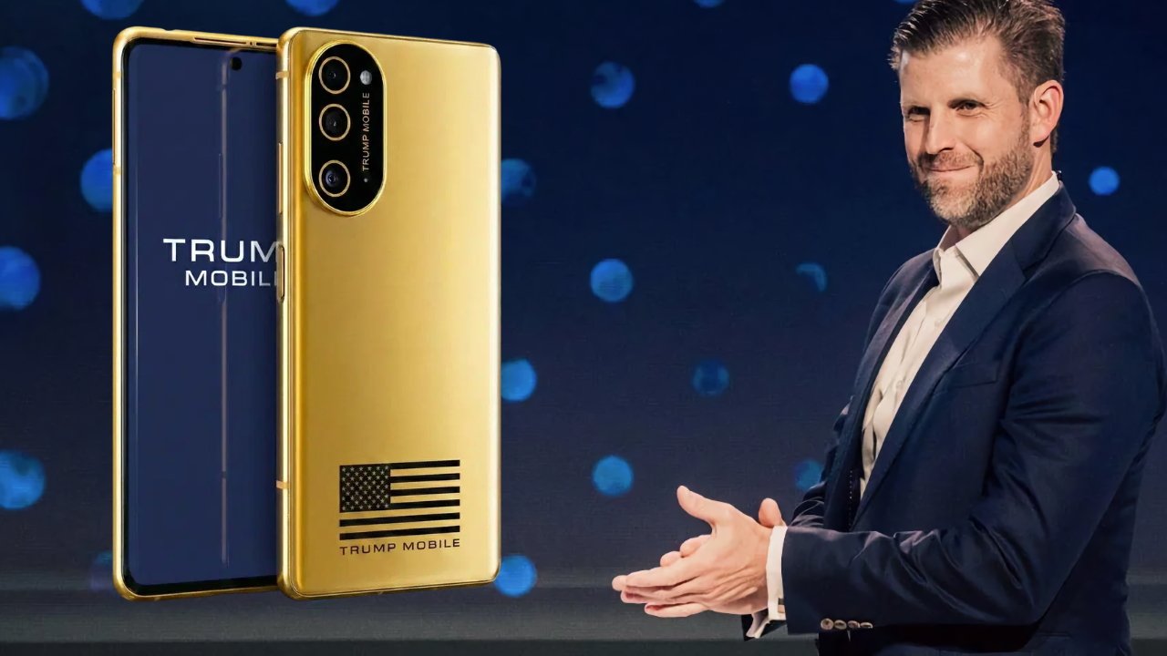 Smiling man in a dark suit claps beside two gold Trump Mobile smartphones, one showing the screen, the other showing rear cameras and an American flag logo on a dark stage background