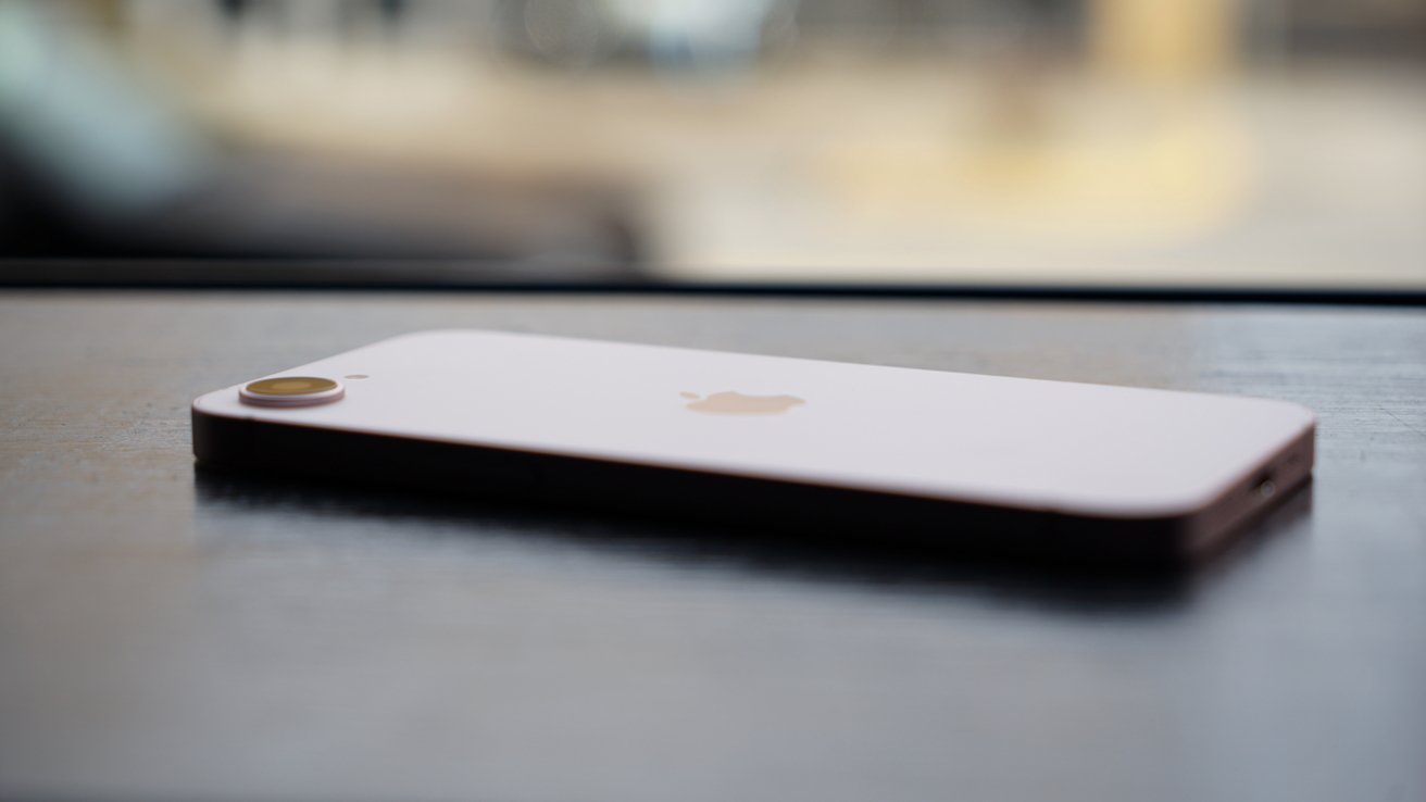 Rose gold iPhone 17e lying face down on a dark table near a window, showing rear camera bump and Apple logo, with softly blurred outdoor background