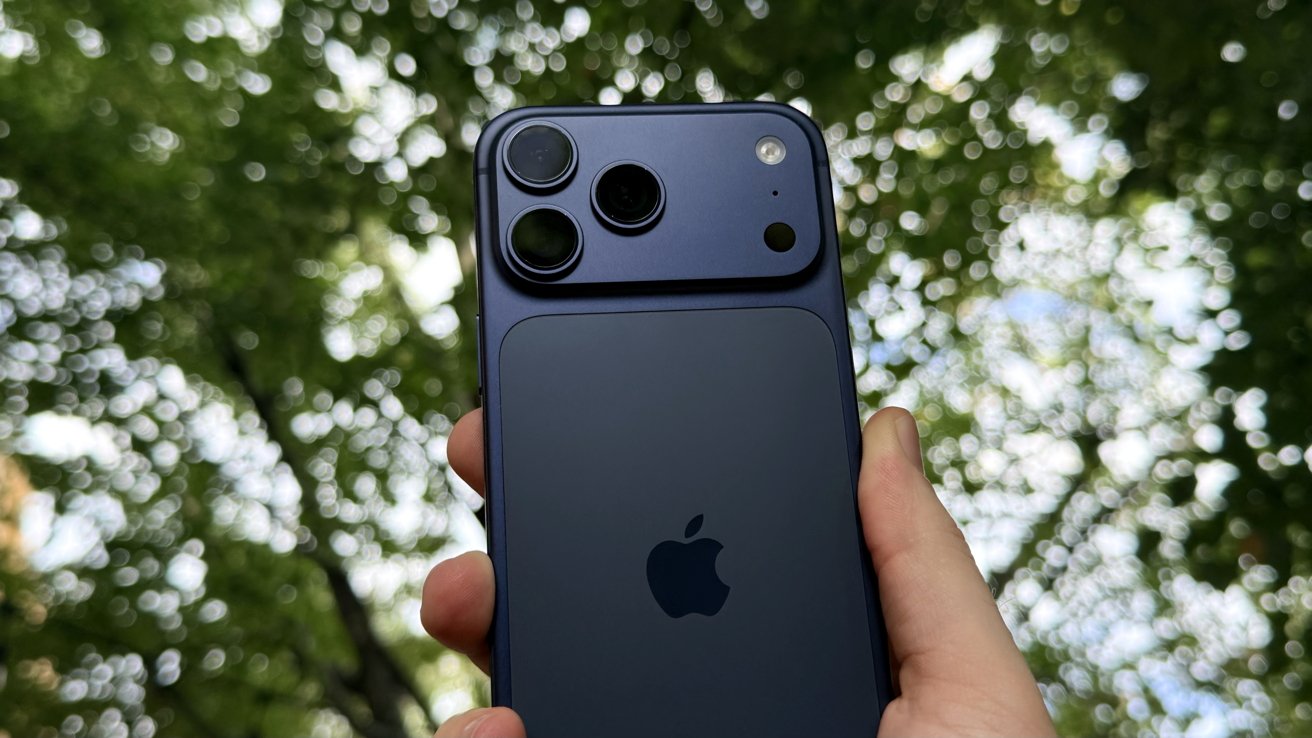 Hand holding a dark blue iPhone 17 Pro Max, focusing on its triple rear cameras and logo, with a softly blurred background of sunlit green tree leaves and bright sky bokeh