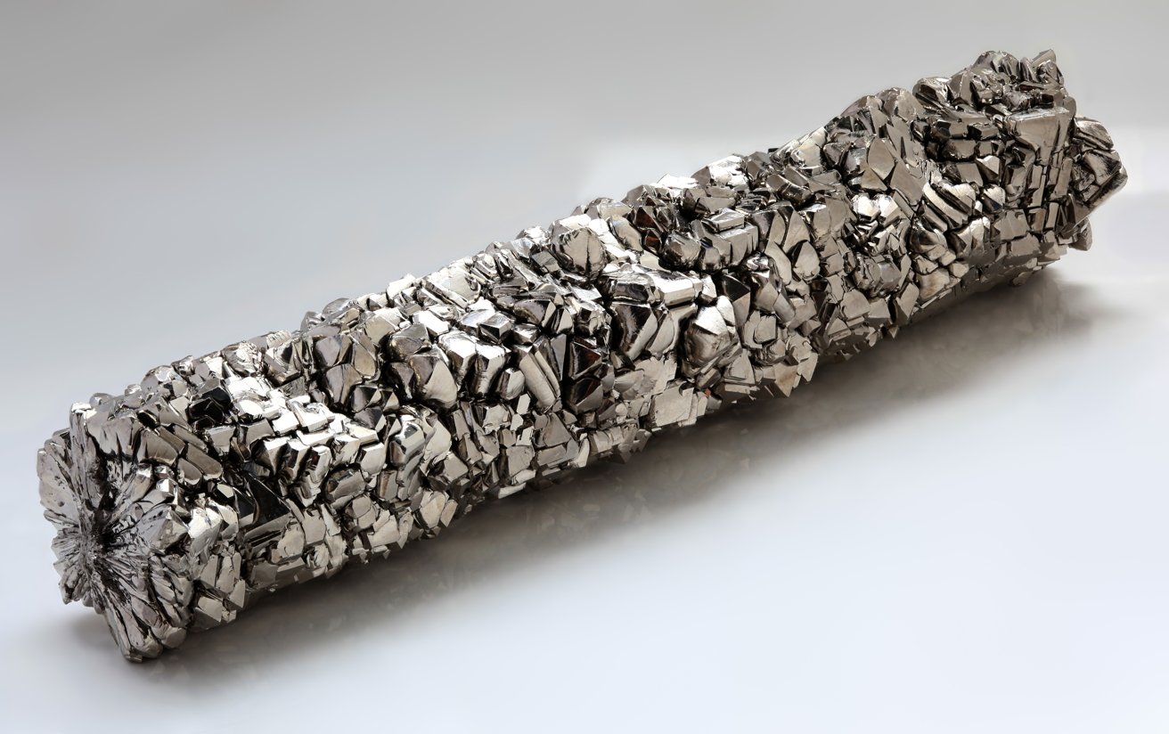 Shiny metallic rod covered in jagged, reflective crystal chunks, resting on a smooth light gray surface with soft shadows
