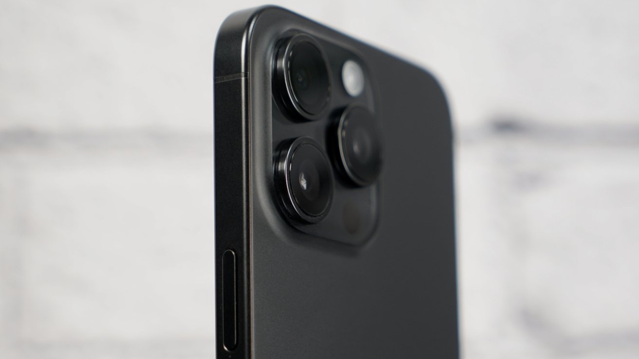 Close-up of an iPhone 15 Pro Max showing three large rear camera lenses and flash on a raised square module against a soft, out-of-focus light background