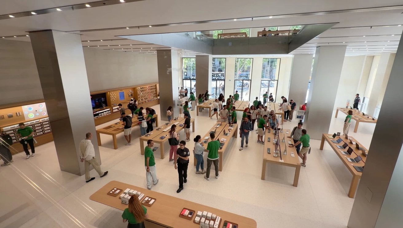 Bright, spacious tech store with wooden tables displaying devices, employees in green shirts assisting numerous customers, large floor-to-ceiling windows, and minimalist white and metal interior design