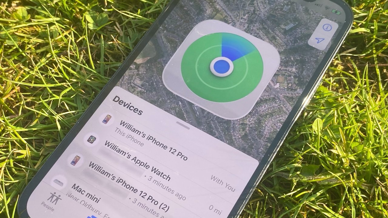 Smartphone lying on green grass, screen showing Apple Find My app with a large location icon and a list of devices including an iPhone, Apple Watch, and Mac mini