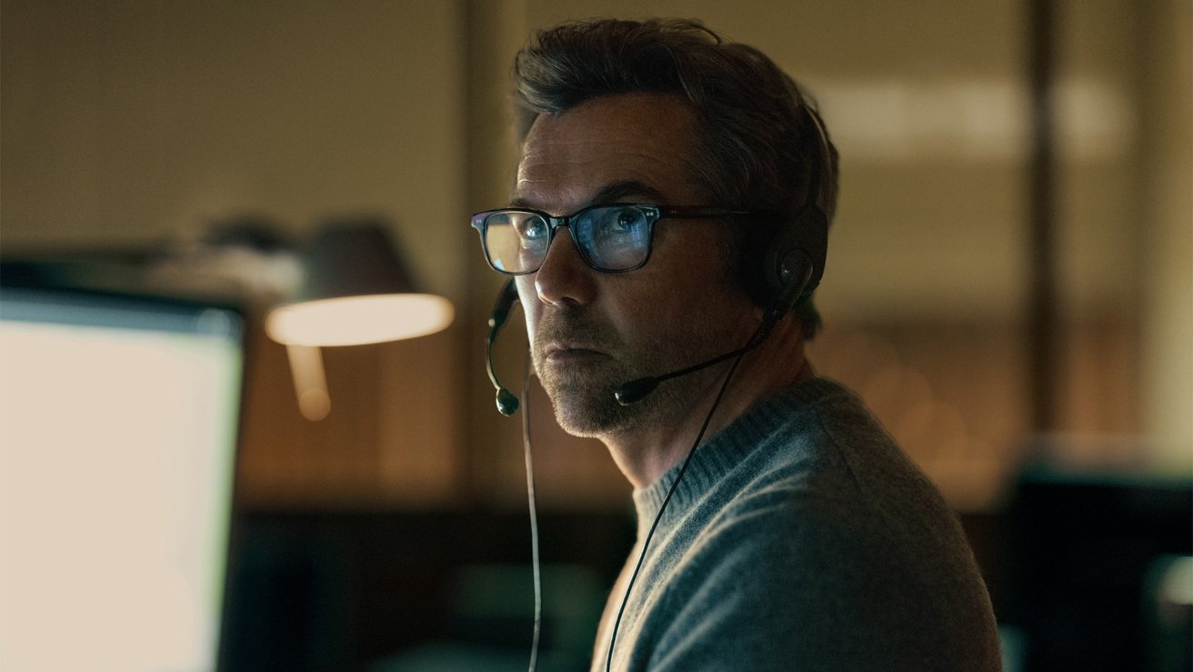Middle-aged man with glasses wearing a headset, sitting at a desk in front of a glowing computer monitor, working intently in a dimly lit office or call center