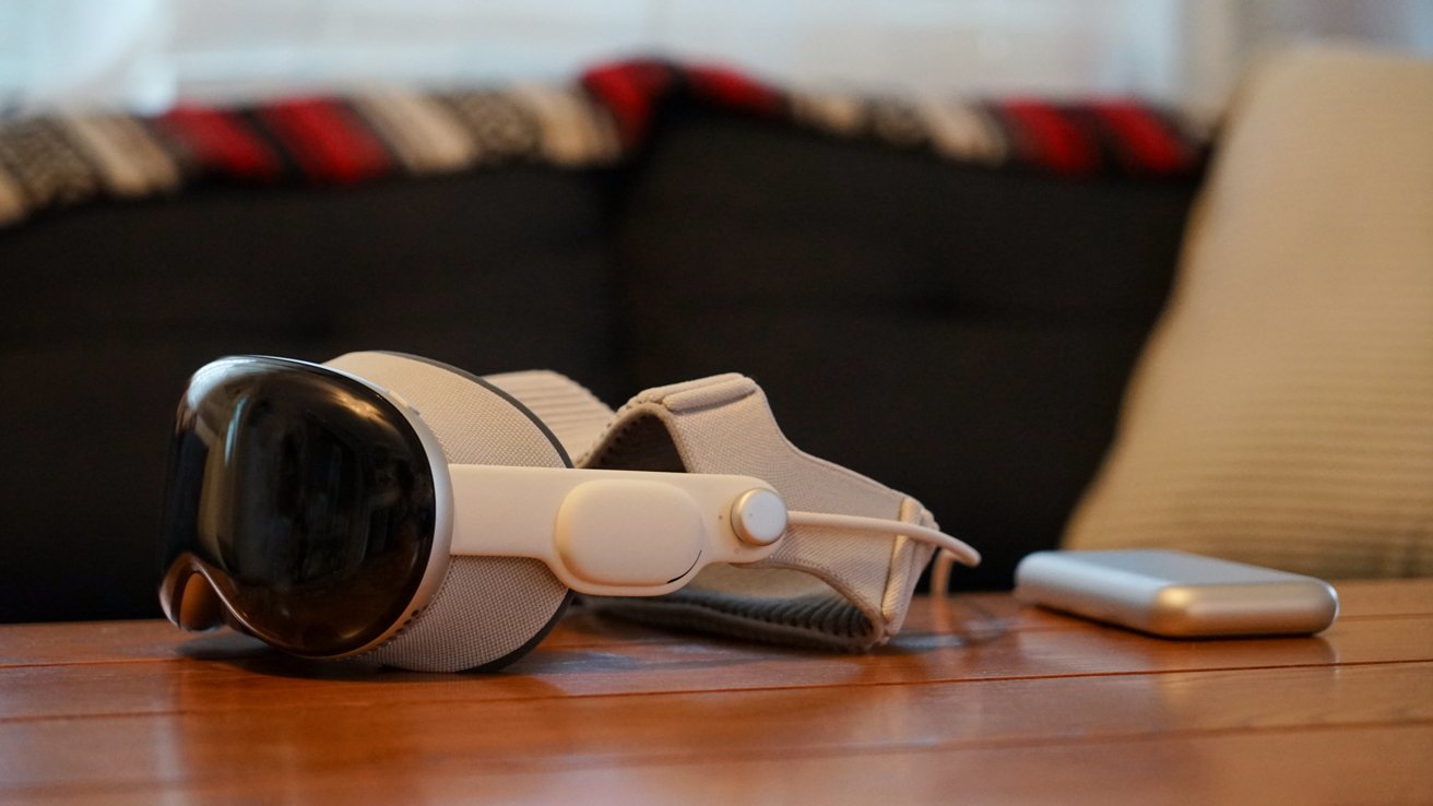 White Apple Vision Pro headset resting on a wooden table beside a small rectangular device, with a dark couch and patterned blanket softly blurred in the background