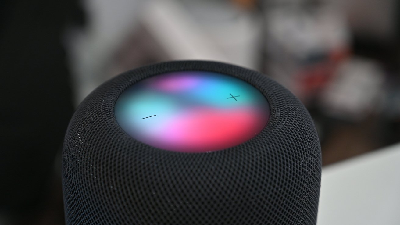 Close-up of a cylindrical smart speaker with mesh fabric and a glowing multicolor touch surface on top, showing plus and minus volume symbols in a blurred indoor setting