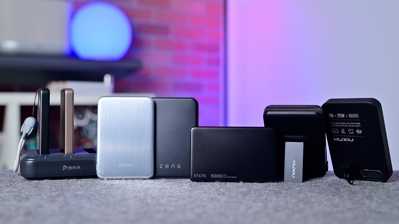 Several portable power banks and charging devices of different sizes and colors arranged in a row on a carpeted surface, with a softly lit brick and purple-blue background