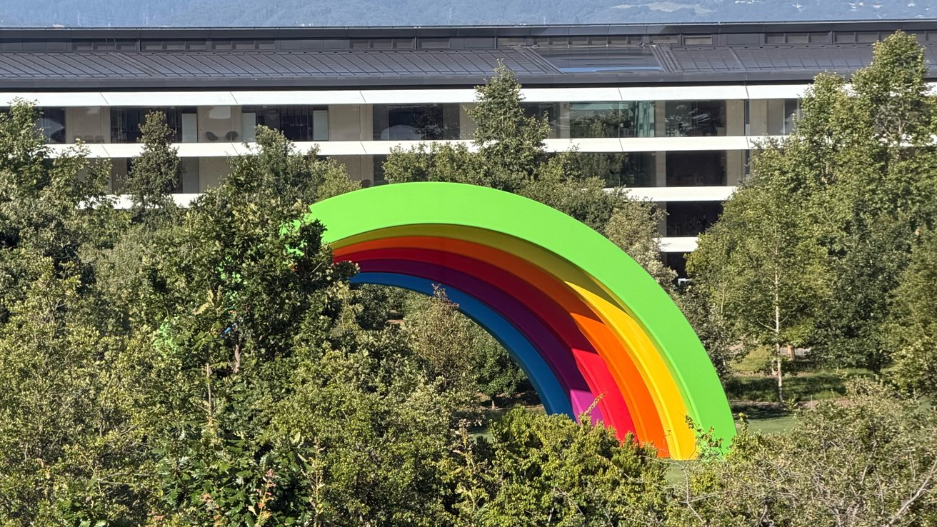 Large colorful rainbow arch sculpture rising above dense green trees, with a modern glass and white office building in the background under a clear sky