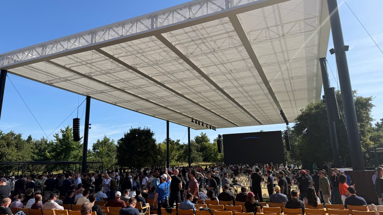 Large outdoor event under a modern white canopy, with rows of wooden seats and many people gathering, facing a large black screen surrounded by trees on a sunny day