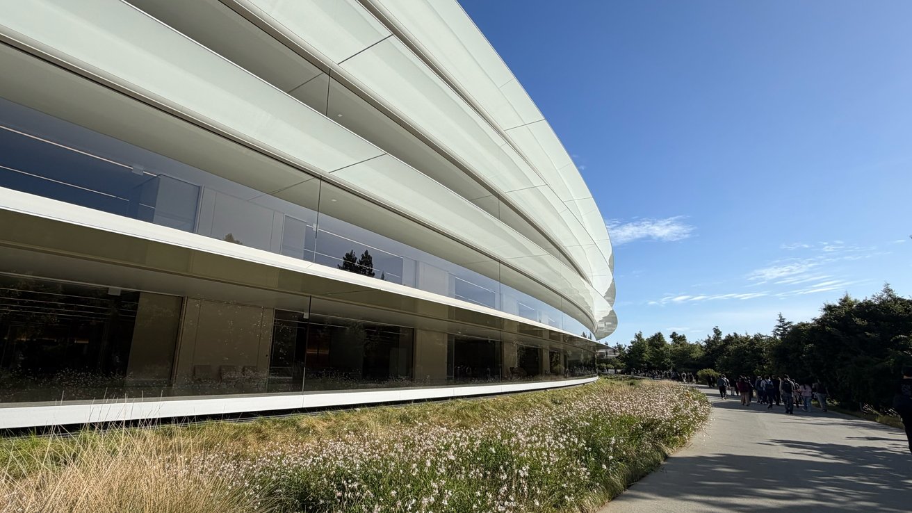 Curved modern glass-and-metal building with reflective windows, bordered by low grasses and flowers, alongside a sunny paved walkway lined with trees under a clear blue sky