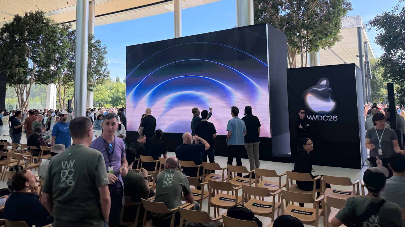 Outdoor Apple WWDC event with attendees gathering around a large screen displaying colorful concentric arcs, surrounded by rows of wooden chairs and tall trees under a modern canopy