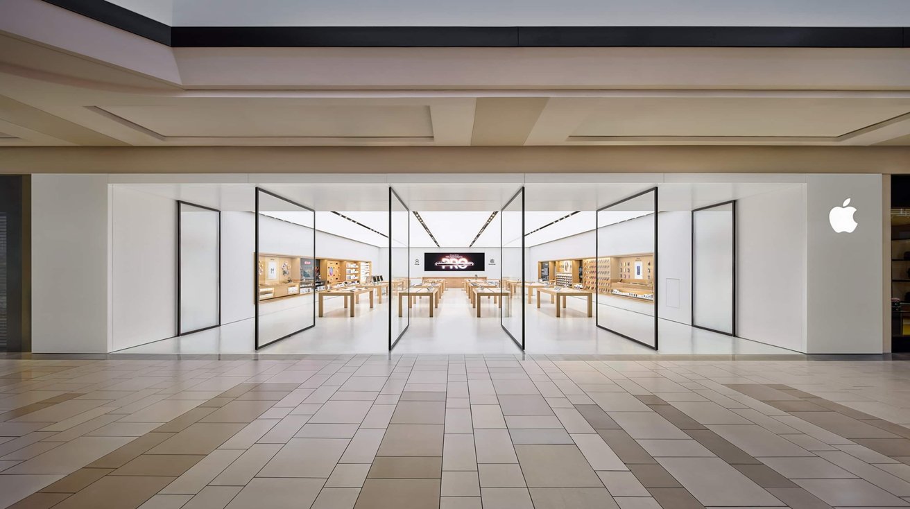 Modern Apple Store in a mall with wide glass entrance, bright white walls, wooden tables displaying devices, large digital screen at the back, and Apple logo on the right wall